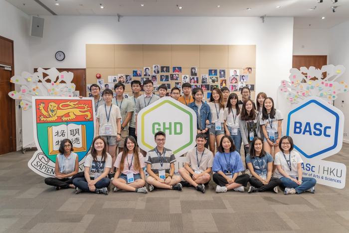 学生坐在前面或站在三个标有 HKU 徽章、GHD 和 BASC 的标志后面